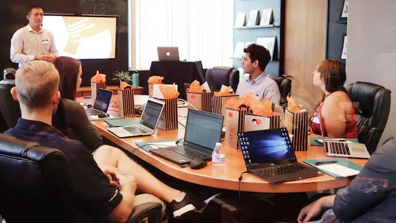A team seated around a boardroom table with laptops, watching a colleague present slides on a wall-mounted screen.