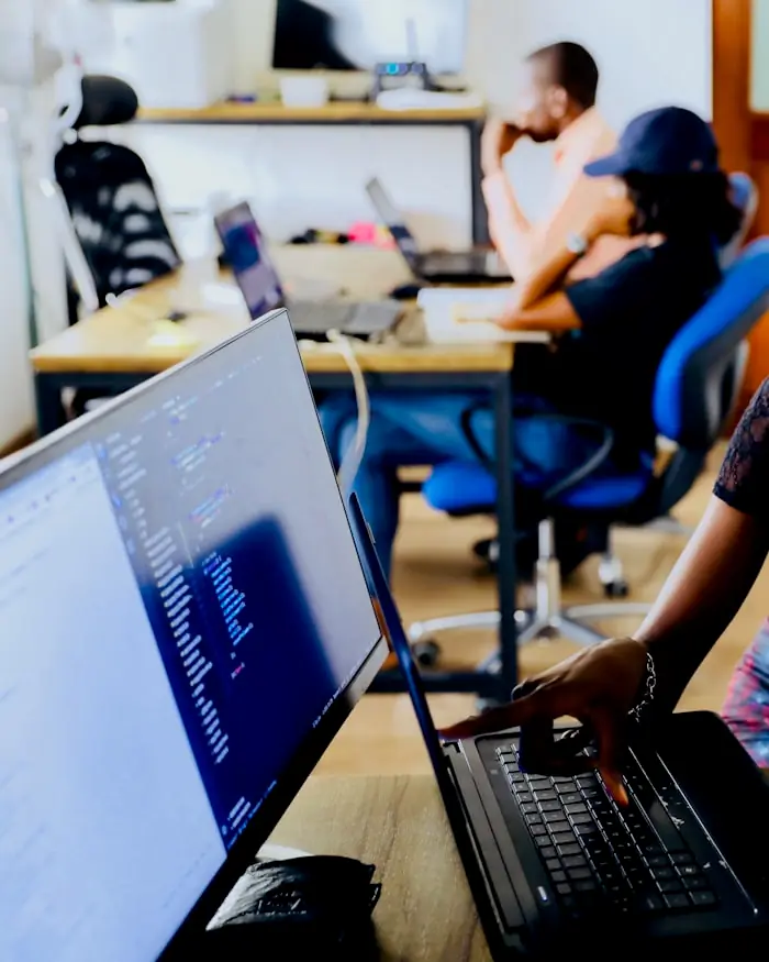 A desk in a shared office with an external monitor showing code, hands typing on a laptop, and colleagues working at desks behind.