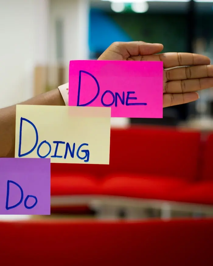 A hand holding three sticky notes reading Do, Doing and Done in marker pen, in front of a red office sofa.