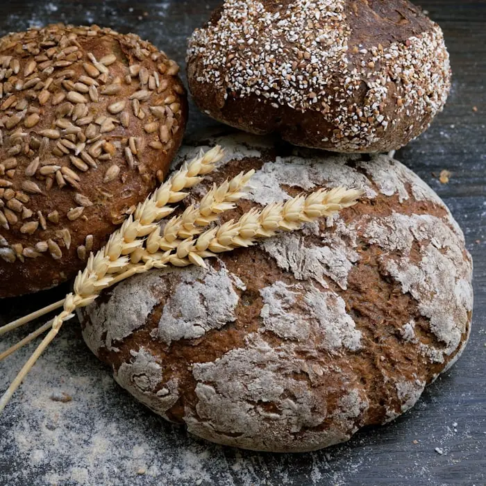 Three round artisan loaves with different seeded crusts, stacked with ears of wheat on a floured dark surface.