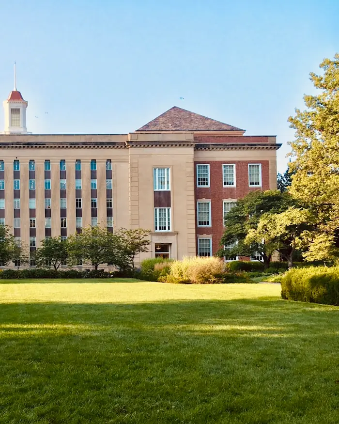 A historic brick and stone college building seen across a sunlit lawn, framed by mature trees.