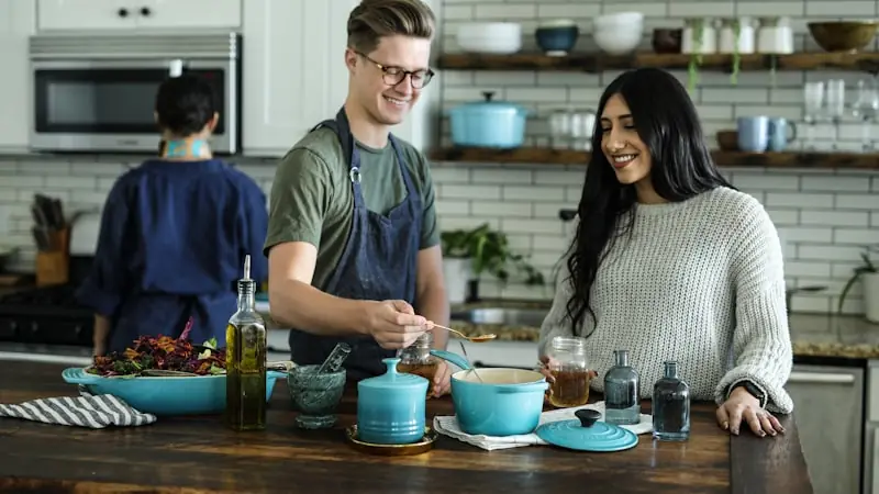Two people in a kitchen, one in an apron spooning from a teal pot on a counter set with oil, glassware and cookware.