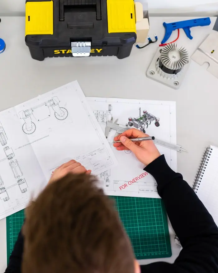 Overhead view of an engineer measuring a small component with digital calipers over a set of technical drawings.