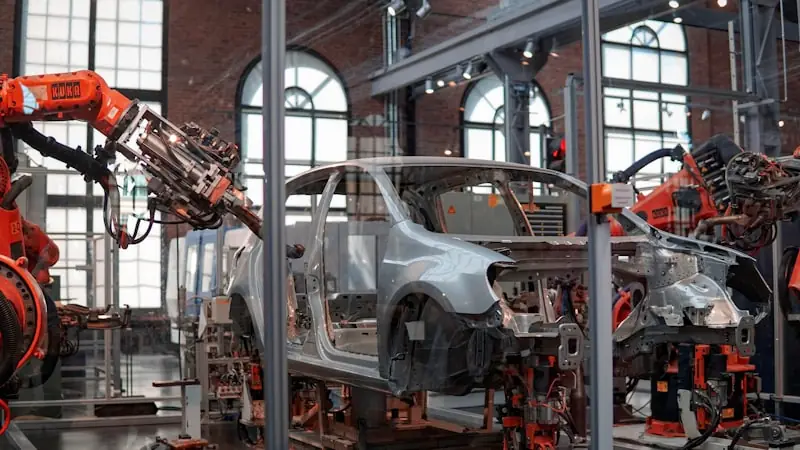 A car body shell moving along an assembly line between orange robotic arms inside a brick-built factory hall.