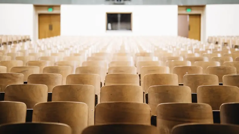 An empty lecture hall photographed from the back, rows of wooden tip-up seats receding towards the front.