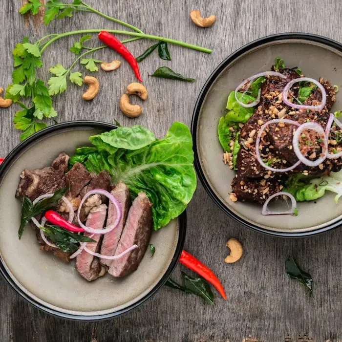 Overhead view of two plates of sliced rare beef with lettuce, red onion, chilli and cashews on a weathered wooden table.