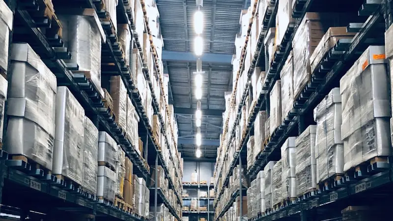 A warehouse aisle between tall racking stacked with shrink-wrapped pallets, lit by a row of overhead lamps.