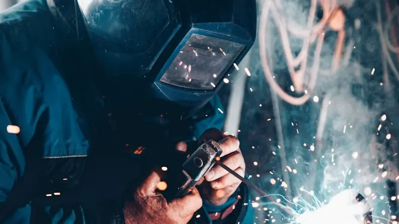 A close-up of a welder in a protective helmet working on a metal pipe, sparks scattering across the frame.