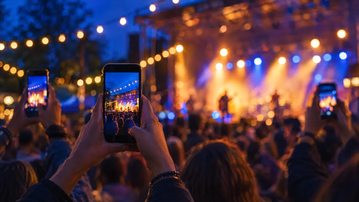 Hands holding up phones at an outdoor evening event, photographing the stage under festoon lights
